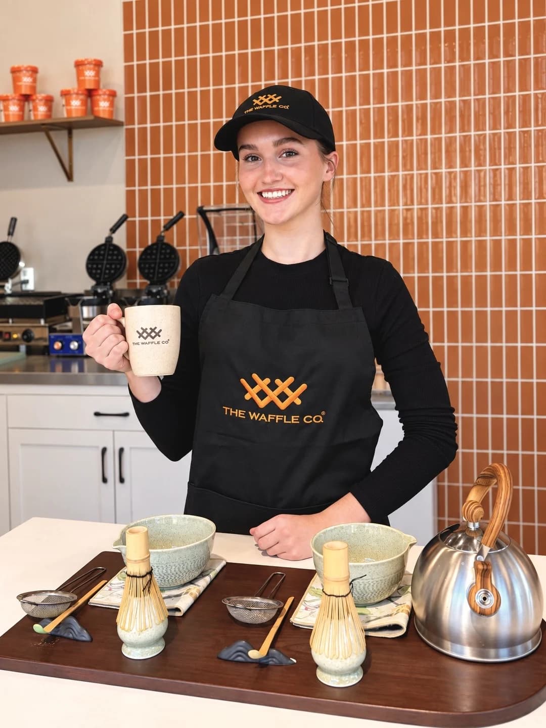 Smiling The Waffle Co. team member in a branded cap and apron, holding a mug at the matcha bar